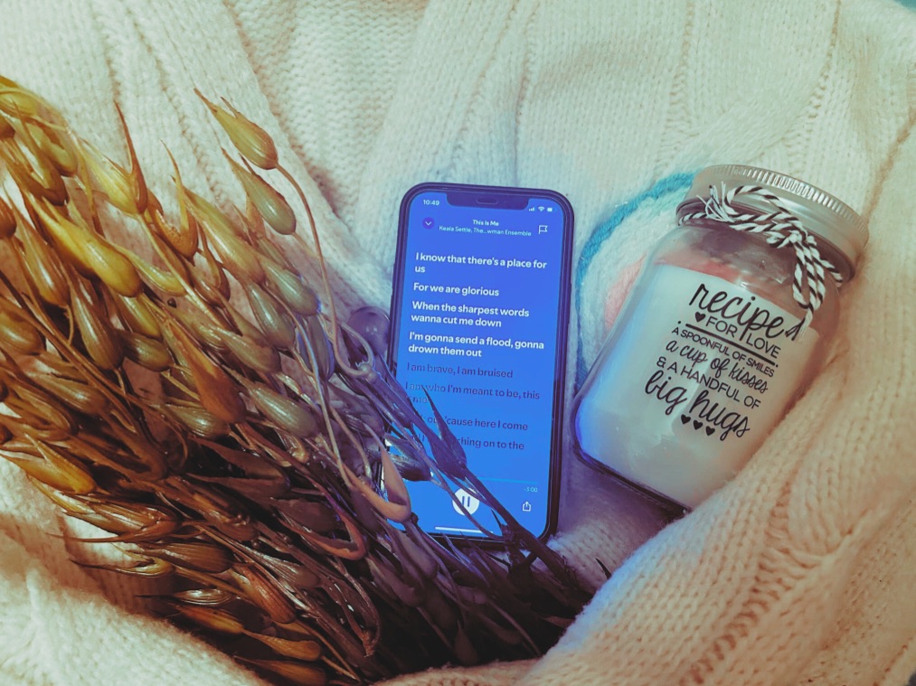 A phone playing “This Is Me”, a faux seaweed plant, a french knitted candle rug and a “Recipe For Love” candle sit within the arms of a cream knitted cardigan.