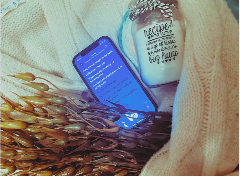 A phone playing “This Is Me”, a faux seaweed plant, a french knitted candle rug and a “Recipe For Love” candle sit within the arms of a cream knitted cardigan.