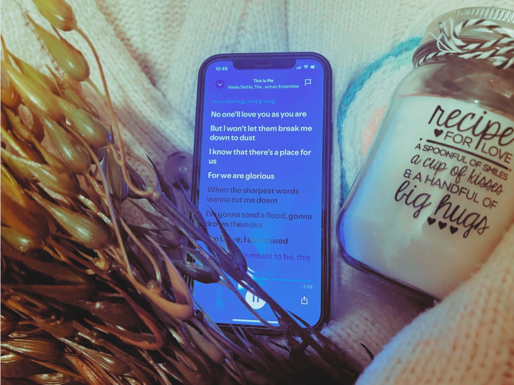 A phone playing “This Is Me”, a faux seaweed plant, a french knitted candle rug and a “Recipe For Love” candle sit within the arms of a cream knitted cardigan.