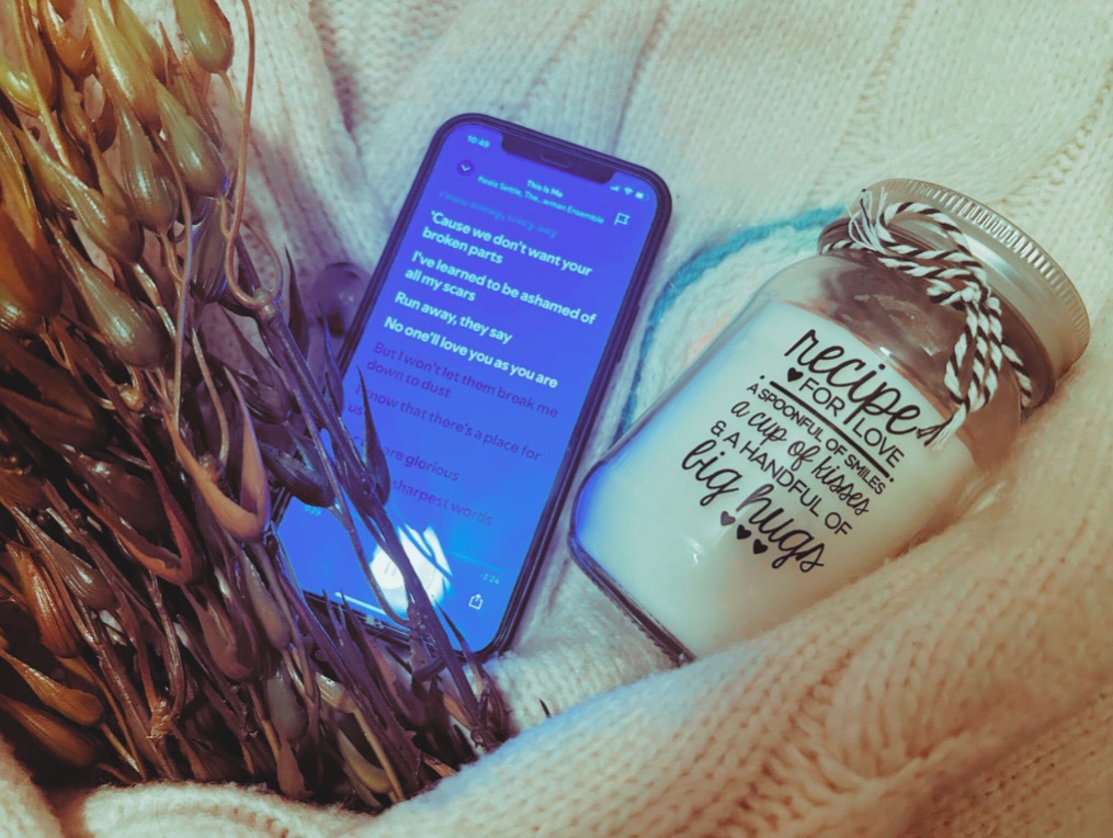 A phone playing “This Is Me”, a faux seaweed plant, a french knitted candle rug and a “Recipe For Love” candle sit within the arms of a cream knitted cardigan.