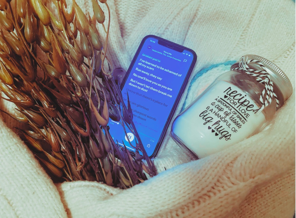 A phone playing “This Is Me”, a faux seaweed plant, a french knitted candle rug and a “Recipe For Love” candle sit within the arms of a cream knitted cardigan.