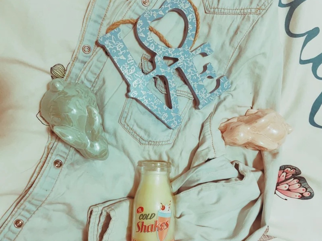 Two pastel ceramic bunnies, a milkshake style candle and a blue wooden ‘love’ sign with a white lace design lay on a denim shirt on a bed.