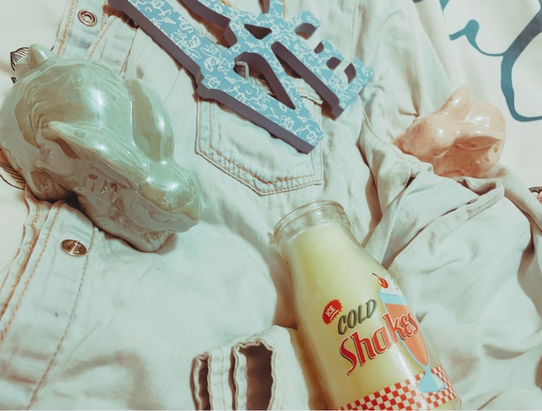 Two pastel ceramic bunnies, a milkshake style candle and a blue wooden ‘love’ sign with a white lace design lay on a denim shirt on a bed.