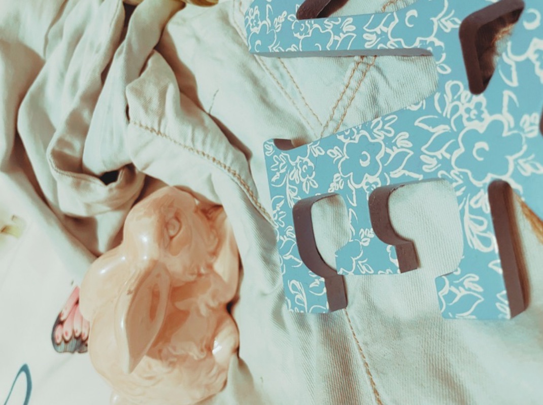 Two pastel ceramic bunnies, a milkshake style candle and a blue wooden ‘love’ sign with a white lace design lay on a denim shirt on a bed.
