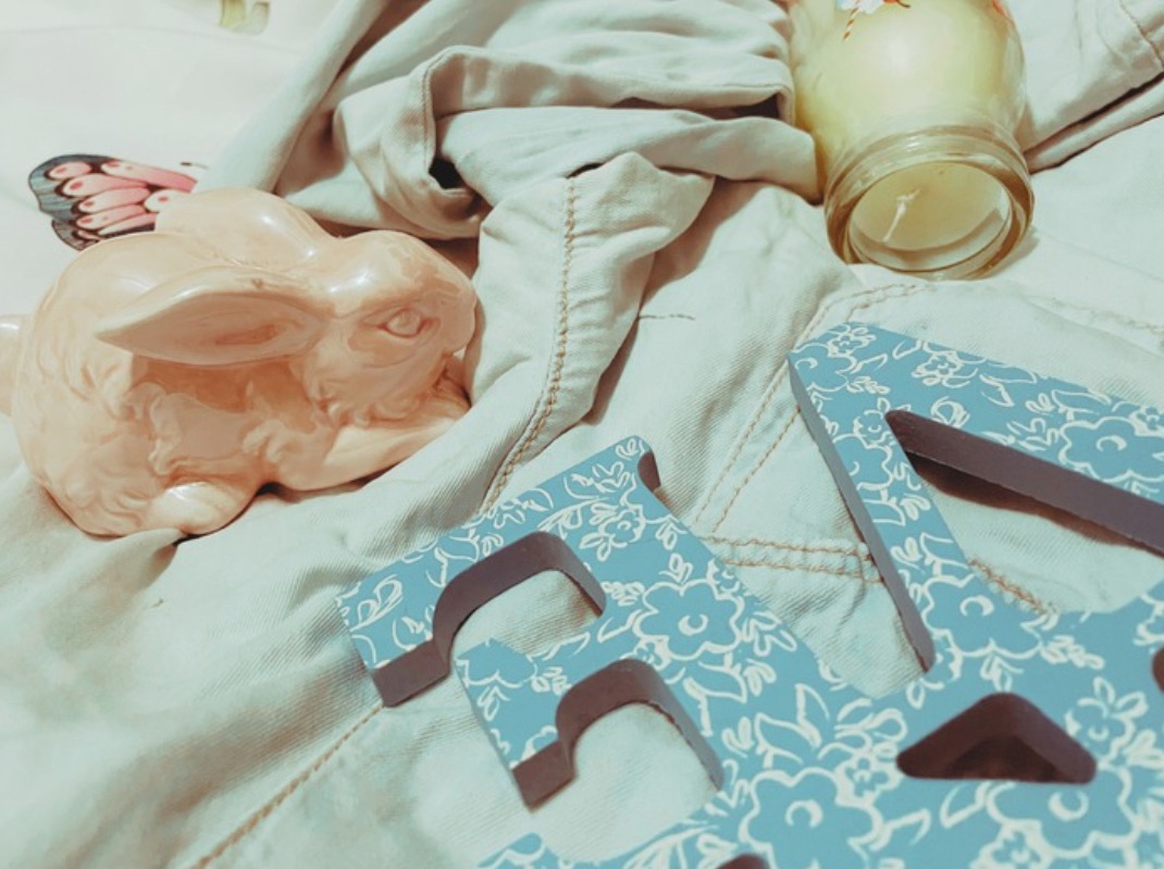 Two pastel ceramic bunnies, a milkshake style candle and a blue wooden ‘love’ sign with a white lace design lay on a denim shirt on a bed.