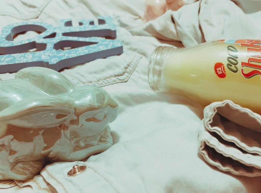 Two pastel ceramic bunnies, a milkshake style candle and a blue wooden ‘love’ sign with a white lace design lay on a denim shirt on a bed.