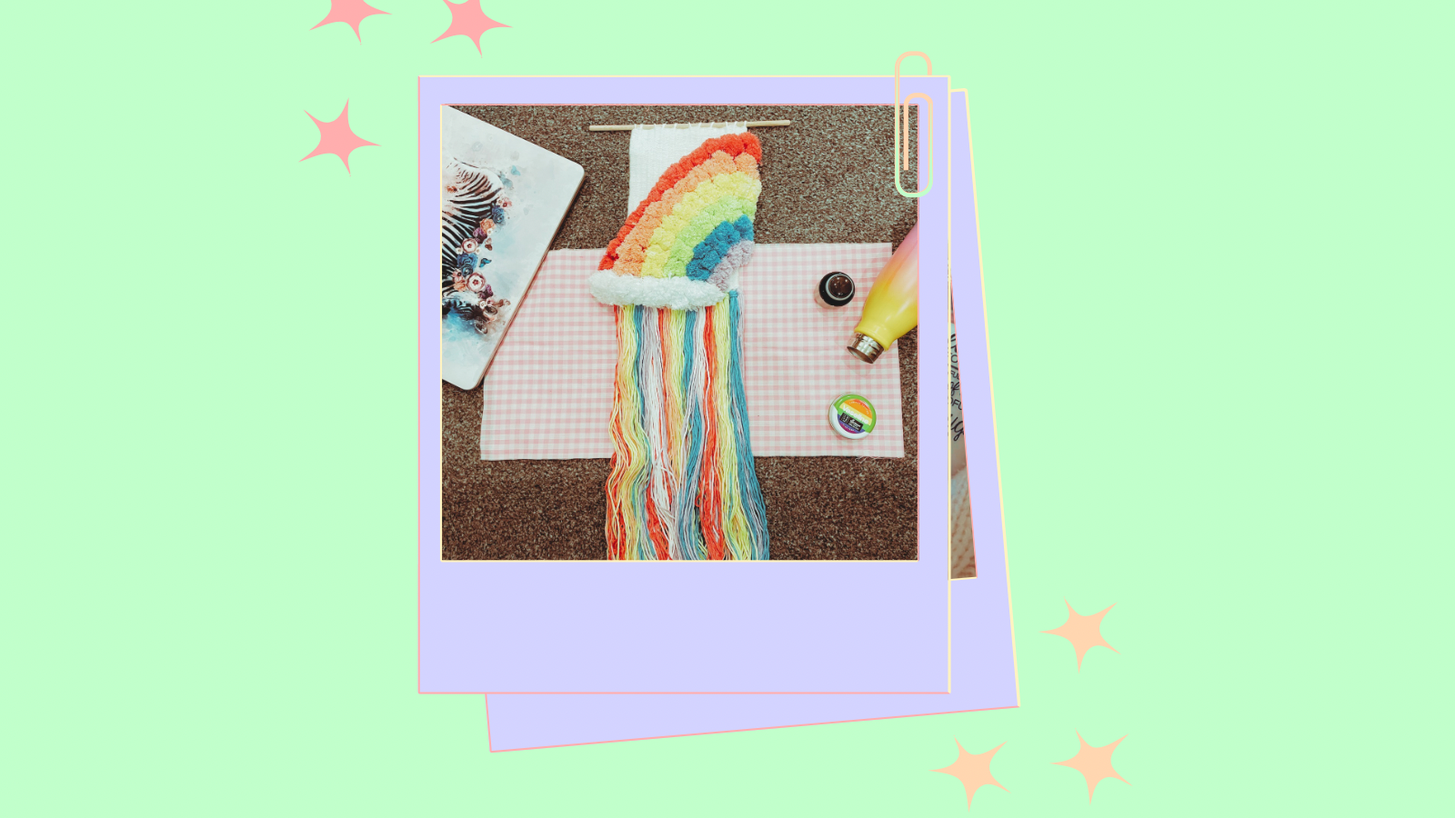 A cartoon style polaroid of a rainbow weave, metal water bottle, Pride vaseline and a laptop on a white and pink gingham fabric. The background features a light orange setting with cartoon stars.