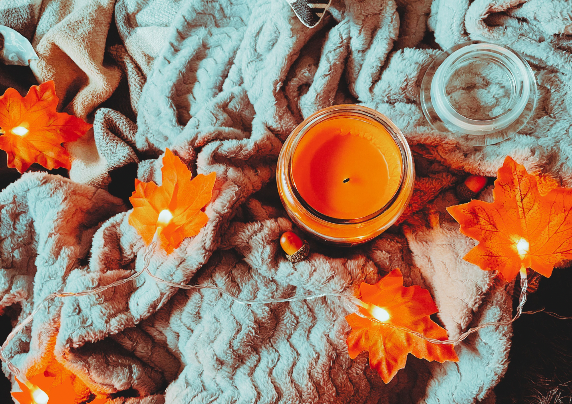 A cozy flat lay featuring a blanket, an autumnal jumper, and autumn decorations, including a pillow, faux leaf fairy lights, a candle, and a small plush pumpkin.