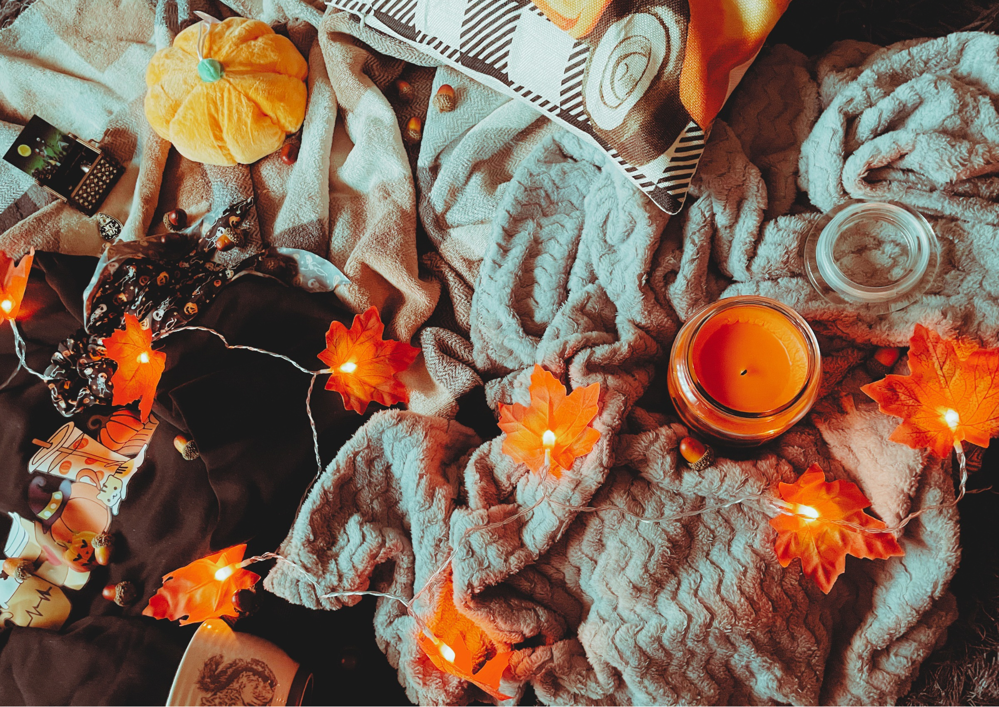 A cozy flat lay featuring a blanket, an autumnal jumper, and autumn decorations, including a pillow, faux leaf fairy lights, a candle, and a small plush pumpkin.