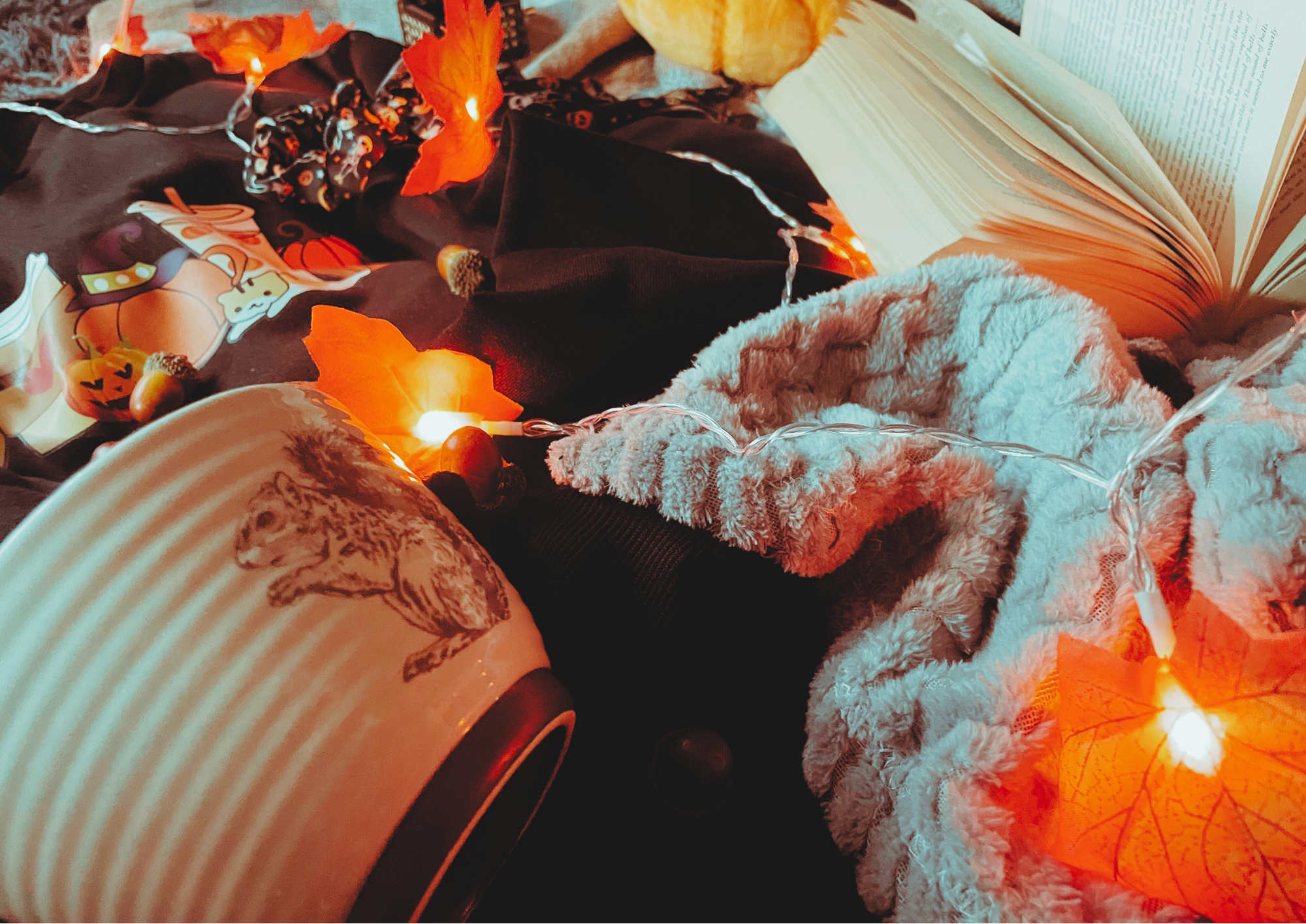 A cozy flat lay featuring a blanket, an autumnal jumper, and autumn decorations, including a pillow, faux leaf fairy lights, a candle, and a small plush pumpkin.