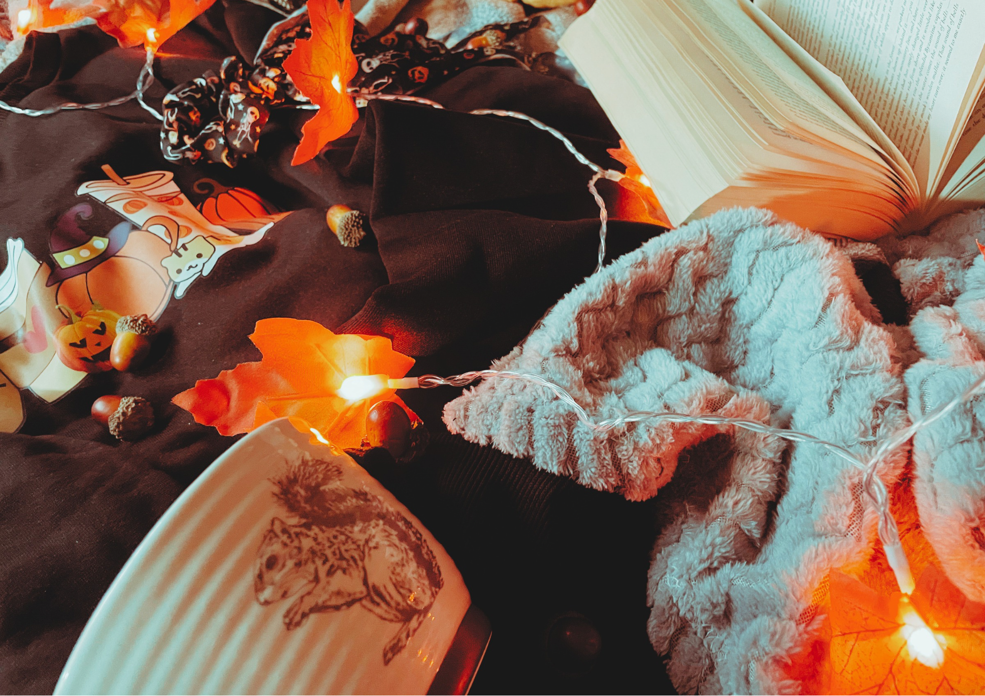 A cozy flat lay featuring a blanket, an autumnal jumper, and autumn decorations, including a pillow, faux leaf fairy lights, a candle, and a small plush pumpkin.