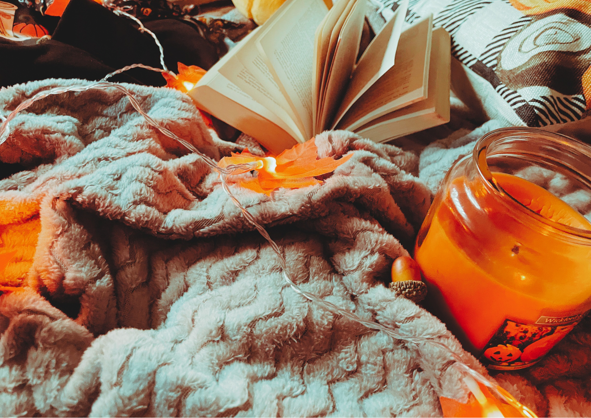 A cozy flat lay featuring a blanket, an autumnal jumper, and autumn decorations, including a pillow, faux leaf fairy lights, a candle, and a small plush pumpkin.