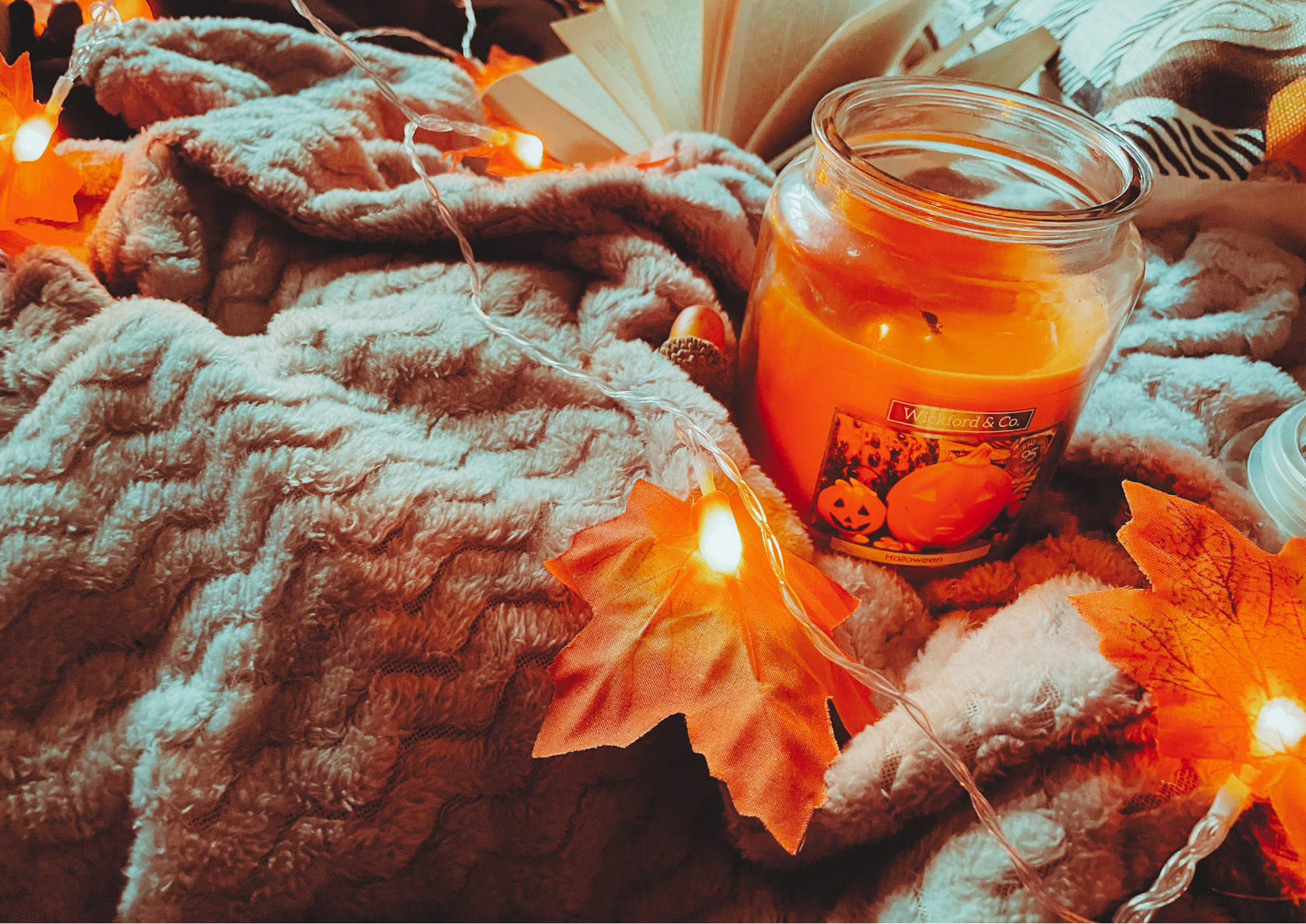 A cozy flat lay featuring a blanket, an autumnal jumper, and autumn decorations, including a pillow, faux leaf fairy lights, a candle, and a small plush pumpkin.