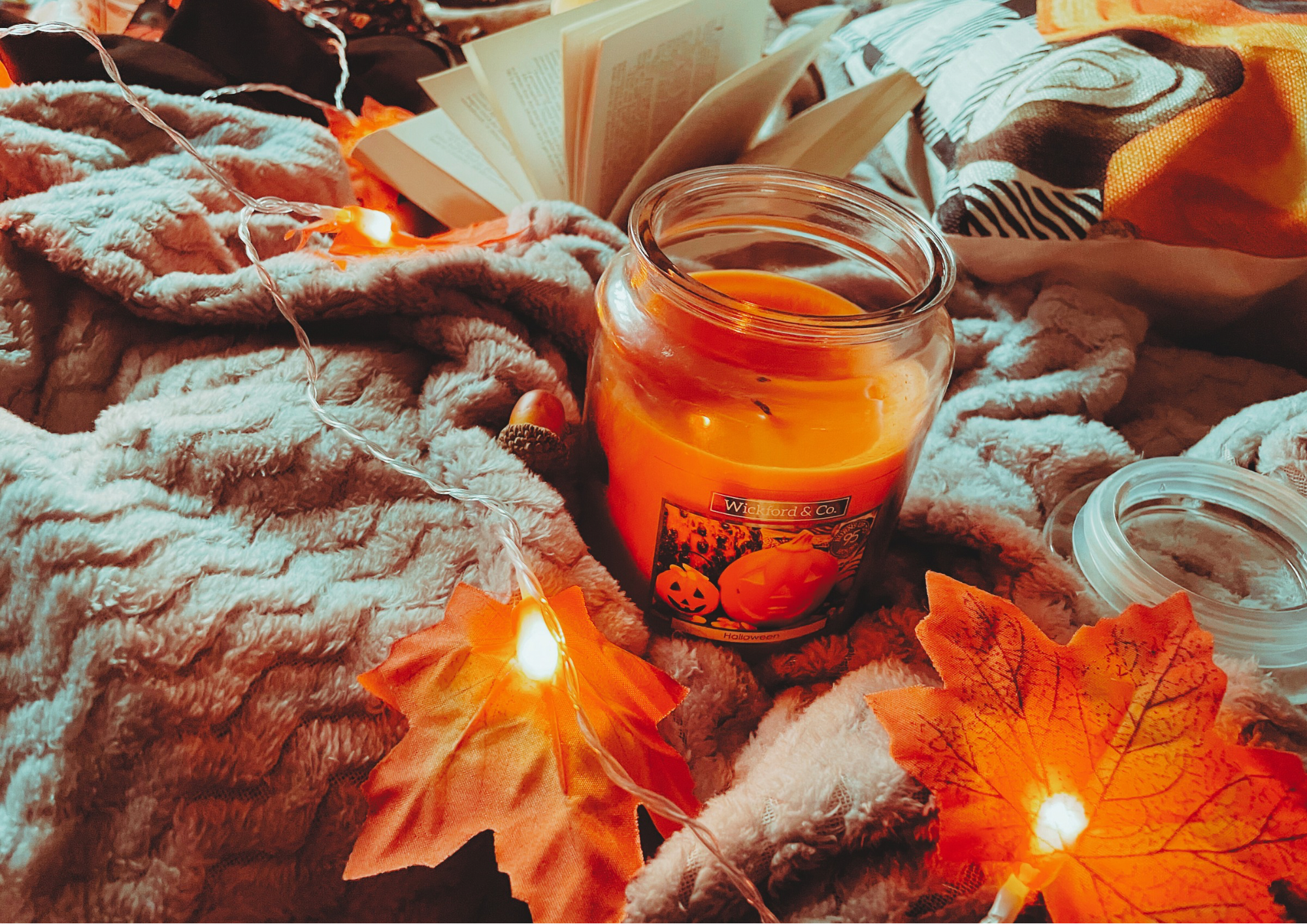 A cozy flat lay featuring a blanket, an autumnal jumper, and autumn decorations, including a pillow, faux leaf fairy lights, a candle, and a small plush pumpkin.