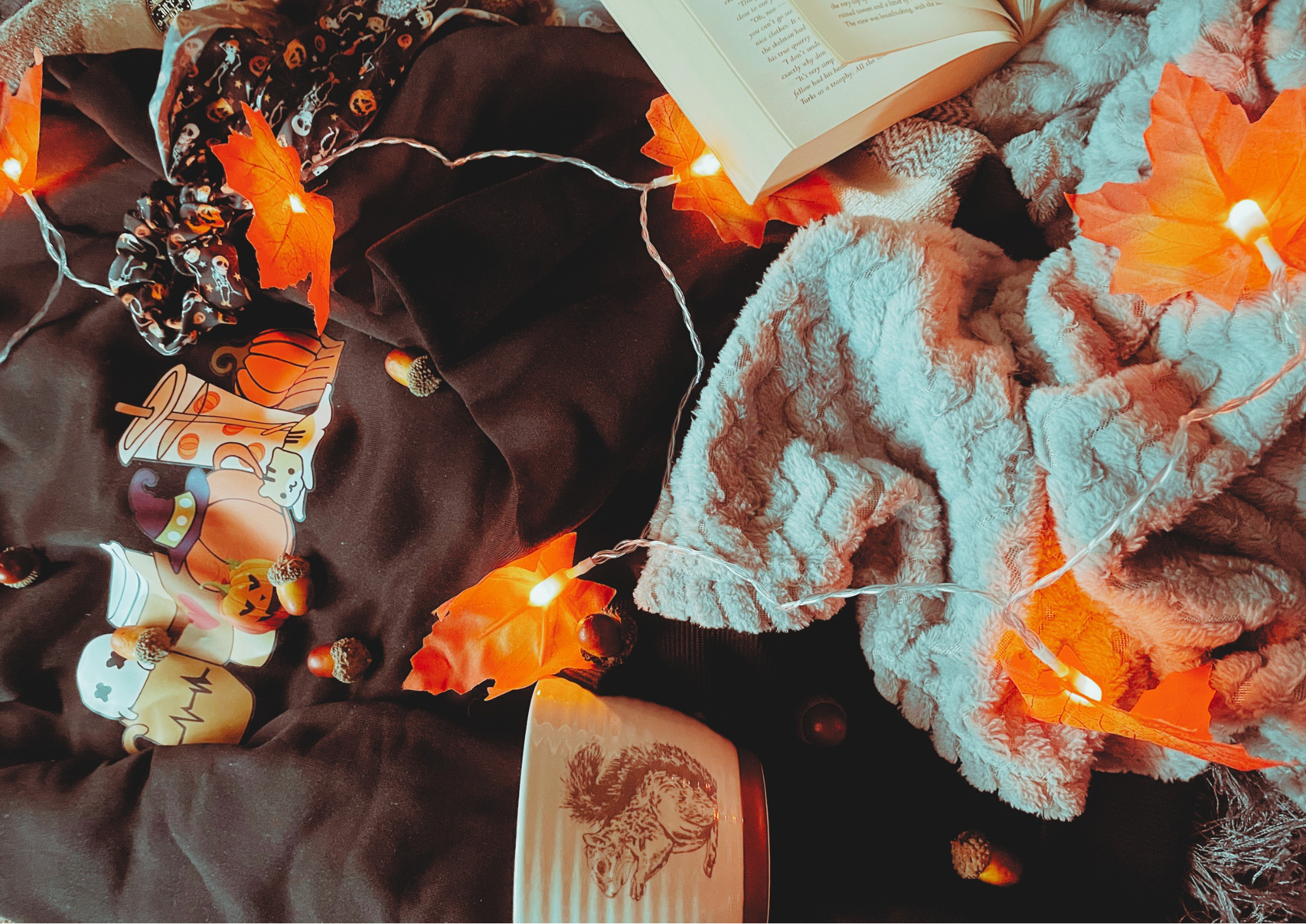 A cozy flat lay featuring a blanket, an autumnal jumper, and autumn decorations, including a pillow, faux leaf fairy lights, a candle, and a small plush pumpkin.