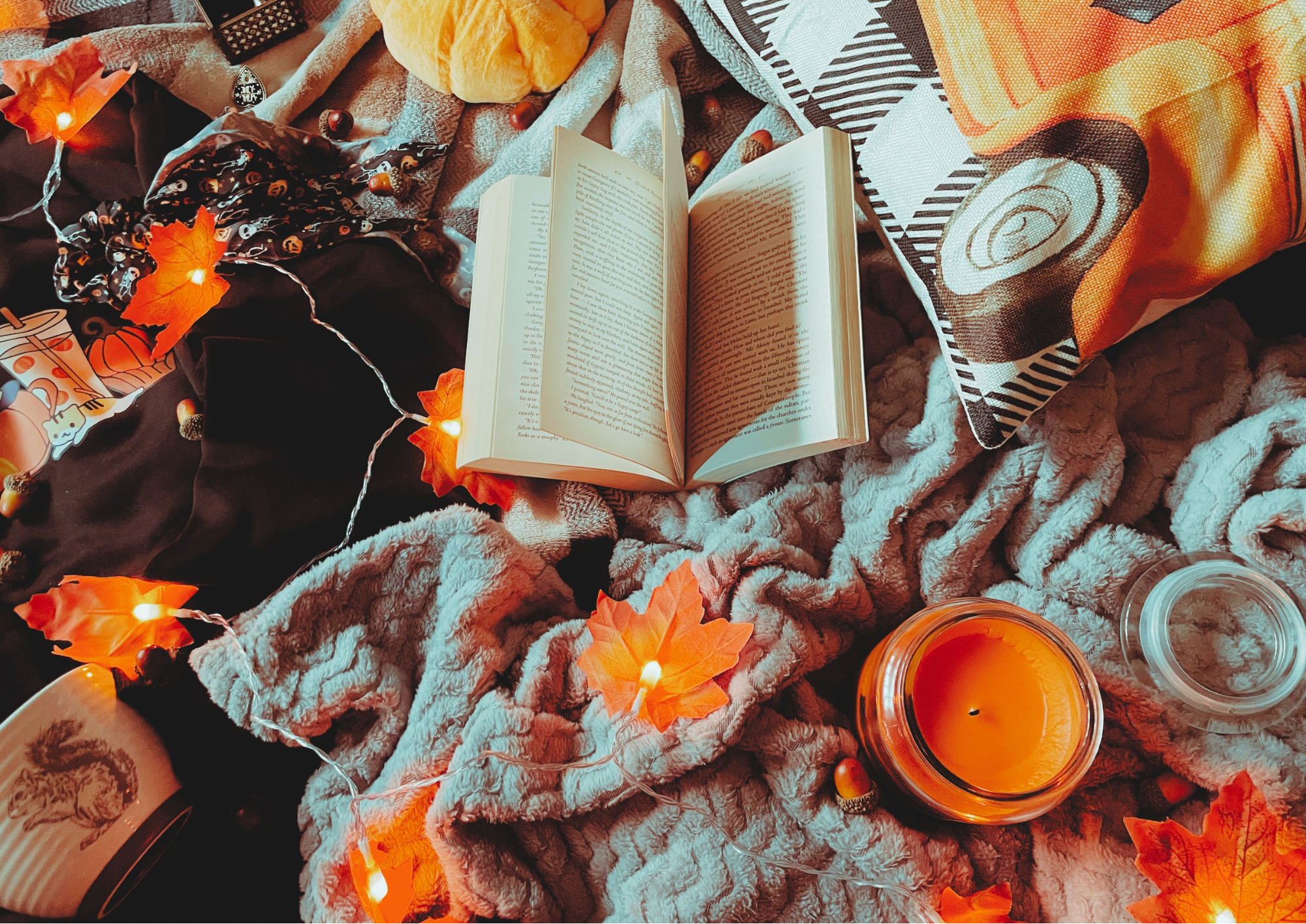 A cozy flat lay featuring a blanket, an autumnal jumper, and autumn decorations, including a pillow, faux leaf fairy lights, a candle, and a small plush pumpkin.